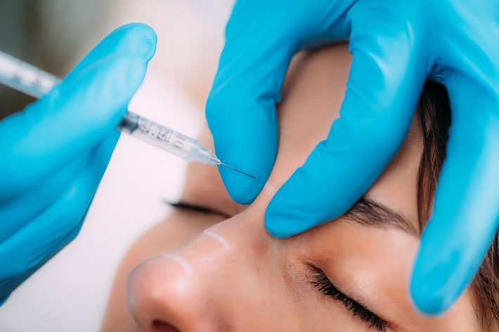 Dermal filler injection treatment. Doctor with surgical gloves injecting dermal filler into women’s nose bridge.