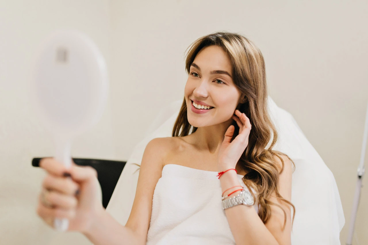 Happy beautiful woman with long brunette hair after cosmetology therapy smiling to mirror in white r