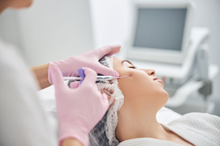 Beauty specialist with gloved hands holding a syringe and applying rejuvenation liquid in female cheek. Facelift