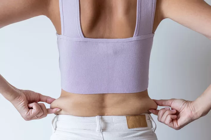 Cropped image of overweight woman, tucking, hiding fat naked big excessive belly with navel in white jeans. Dangling down stomach, big size tummy. Drag away of abdomen. Go on diet, liposuction.